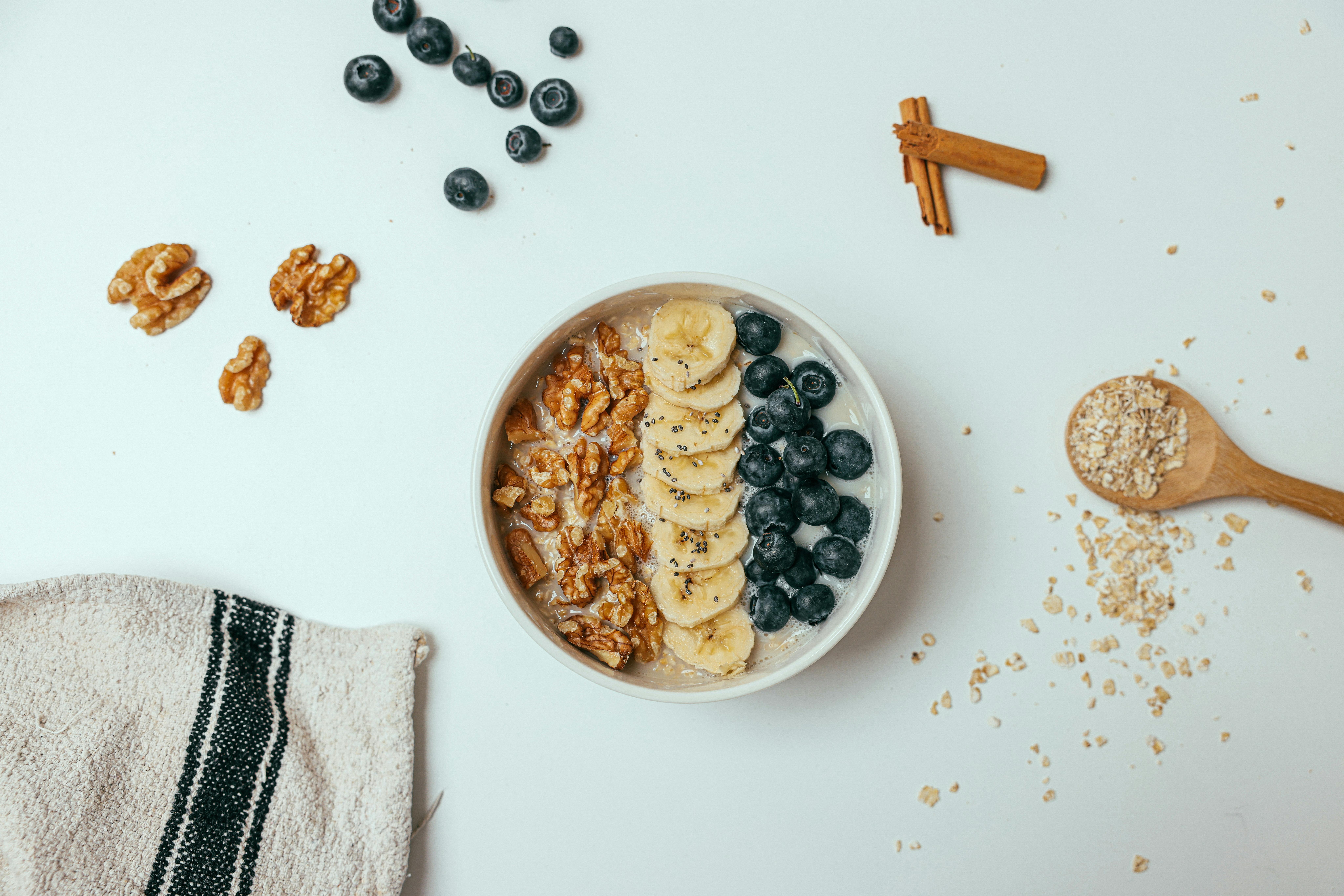 A delicious and healthy breakfast bowl topped with bananas, blueberries, and walnuts on a white surface.